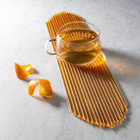 A glass cup of tea sits on a golden honeycomb-textured mat, flanked by orange peels—the perfect scene to sweeten your drink with Orange Blossom Honey Straws and enjoy alongside healthy snacks.