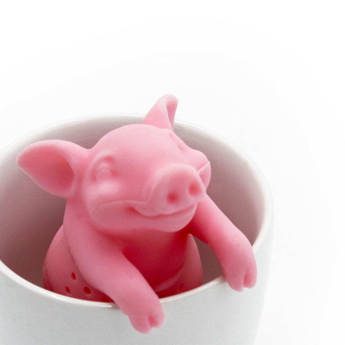 The Hot Belly Infuser, a pink pig-shaped tea infuser made from BPA-free silicone, sits inside a white cup with its front legs hanging over the rim and a cheerful face. The background is plain white.