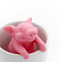 The Hot Belly Infuser, a pink pig-shaped tea infuser made from BPA-free silicone, sits inside a white cup with its front legs hanging over the rim and a cheerful face. The background is plain white.