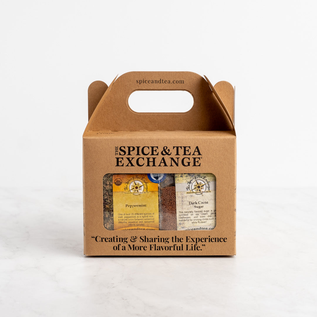 The Hot Mess De-Stress Spa Gift Box from The Spice & Tea Exchange features a brown cardboard box with a handle, clear window, and relaxing Peppermint and Dark Cocoa teas inside. The box reads: "Creating & Sharing the Experience of a More Flavorful Life.