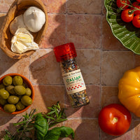A bottle of Italian Street Fair Spice Blend rests on a tiled surface, surrounded by garlic, green olives, fresh basil, a yellow bell pepper, red and cherry tomatoes on a green plate, with bright sunlight casting inviting shadows.