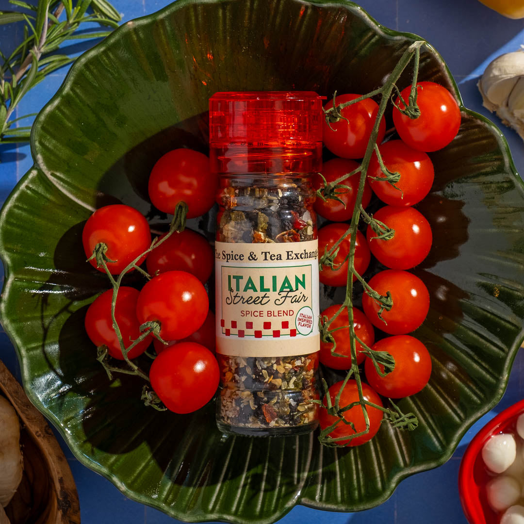 A green leaf-shaped dish holds a bottle of Italian Street Fair Spice Blend, surrounded by vine-ripened cherry tomatoes, with garlic and herbs scattered nearby on a blue surface.