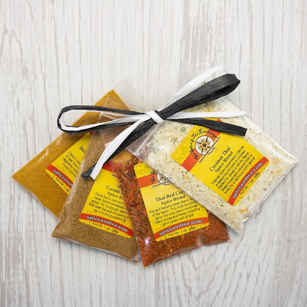 The Keep Calm & Curry On Sampler features three colorful spice blend packets with yellow and red labels, arranged on a light wooden surface and tied with a black and white ribbon for a neat gift bundle.