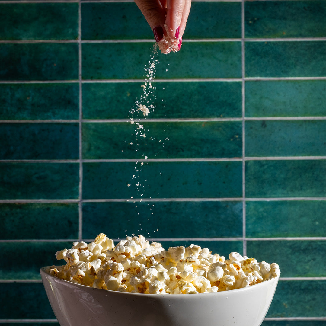 Person sprinkling sugar over popcorn in a bowl against a teal tiled wall.