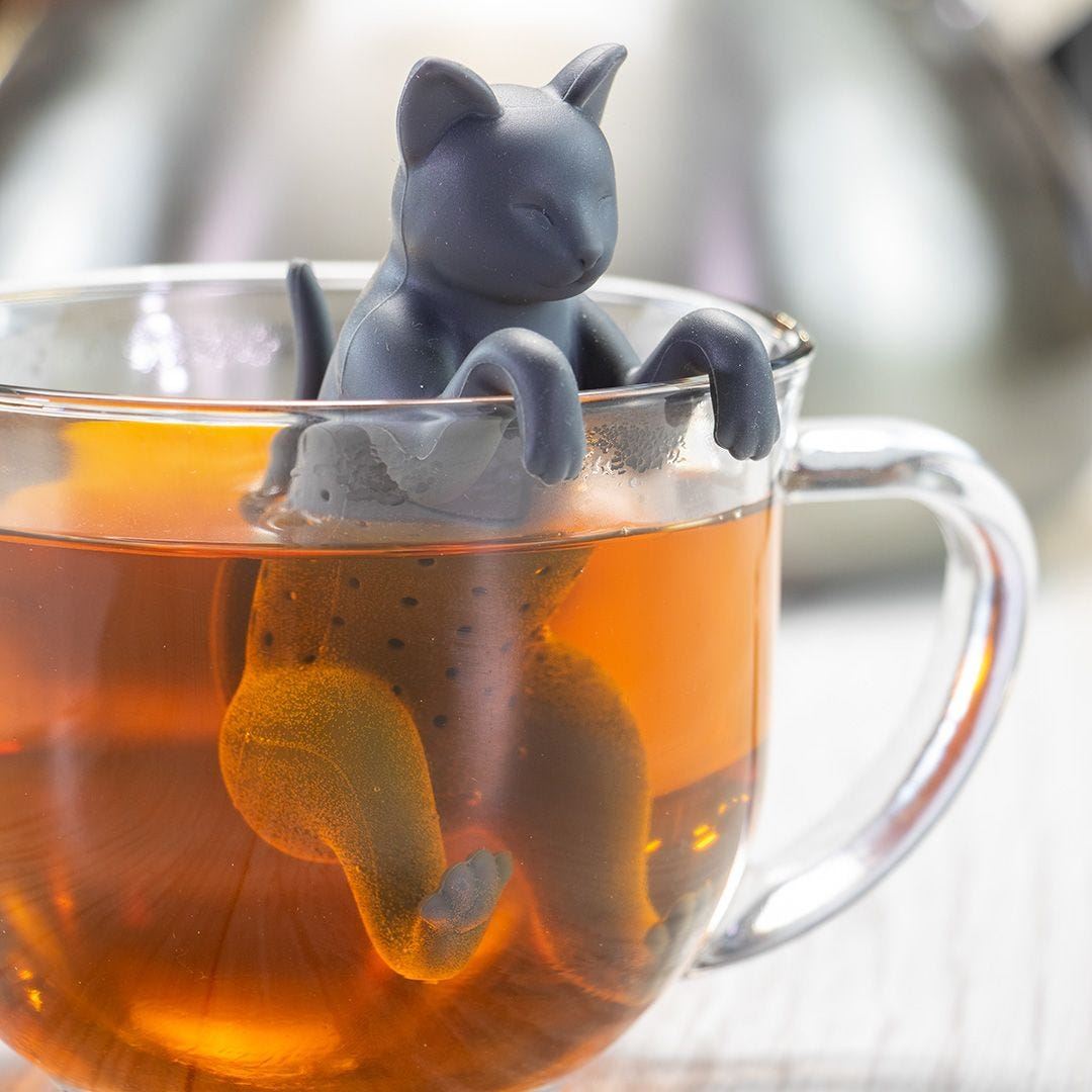 A close-up of a glass cup filled with tea features the Cat Infuser, a BPA-free silicone kitty with its head and paws resting on the rim while its body steeps in the tea.