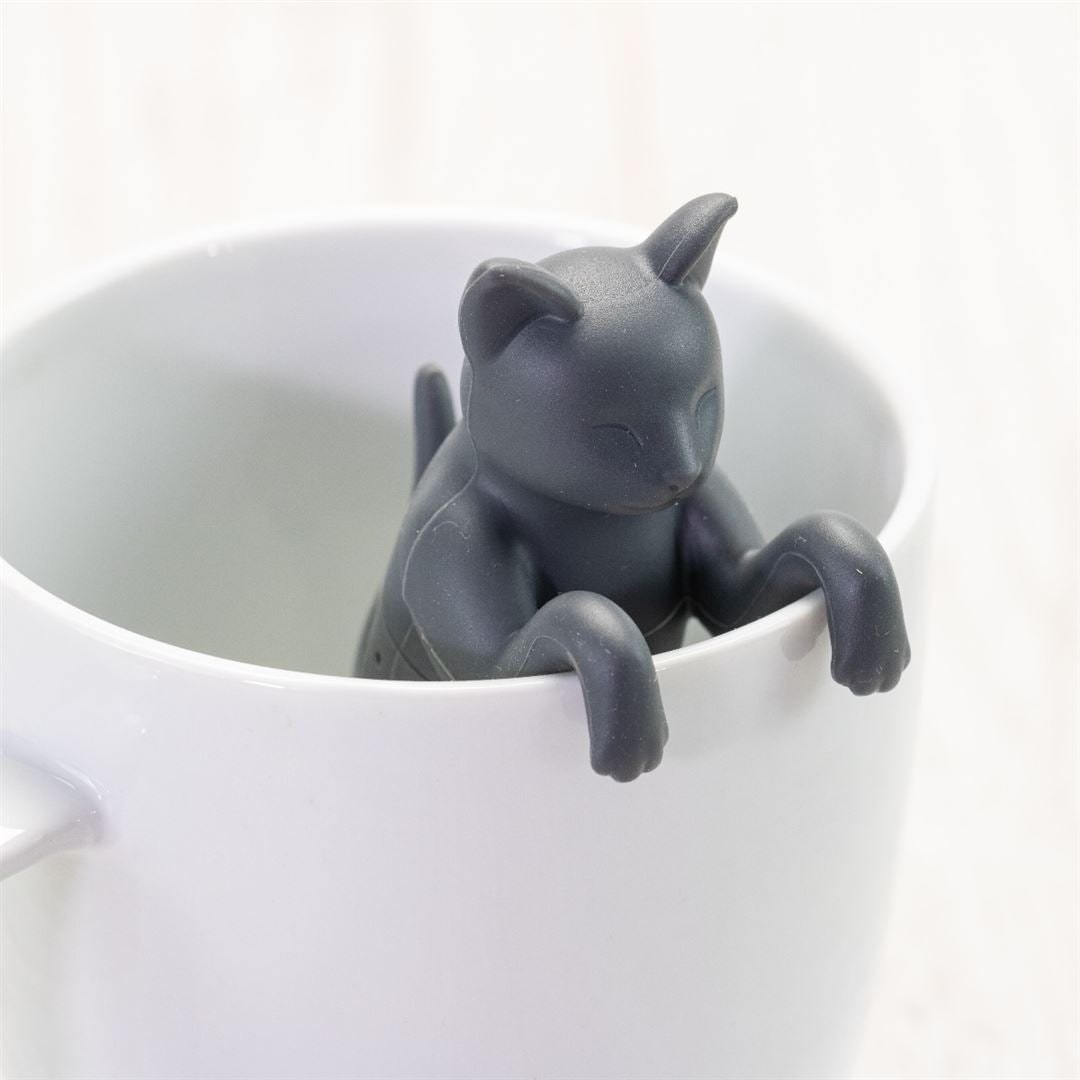The Cat Infuser, a small dark gray kitty made from BPA-free silicone, hangs on the rim of a white ceramic mug with its front paws resting on the edge.