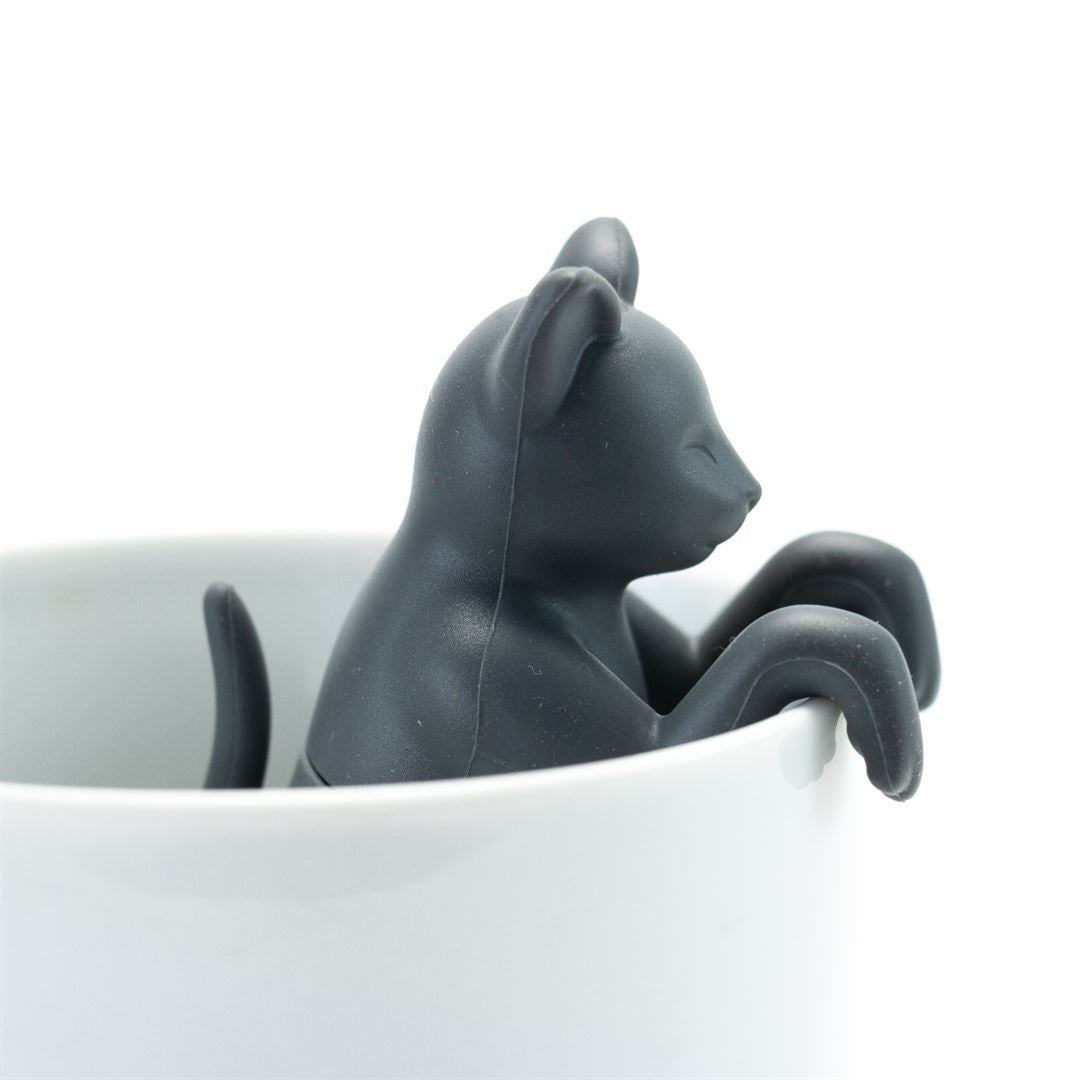 The Cat Infuser, a black cat-shaped tea infuser made from BPA-free silicone, sits on the rim of a white mug with its front paws hanging over the edge.