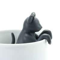 The Cat Infuser, a black cat-shaped tea infuser made from BPA-free silicone, sits on the rim of a white mug with its front paws hanging over the edge.