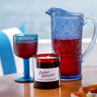 A Lychee Hibiscus Candle, with its tropical scent, sits on a wooden table beside a blue glass goblet and matching blue pitcher filled with a red drink, set against a softly blurred outdoor backdrop.