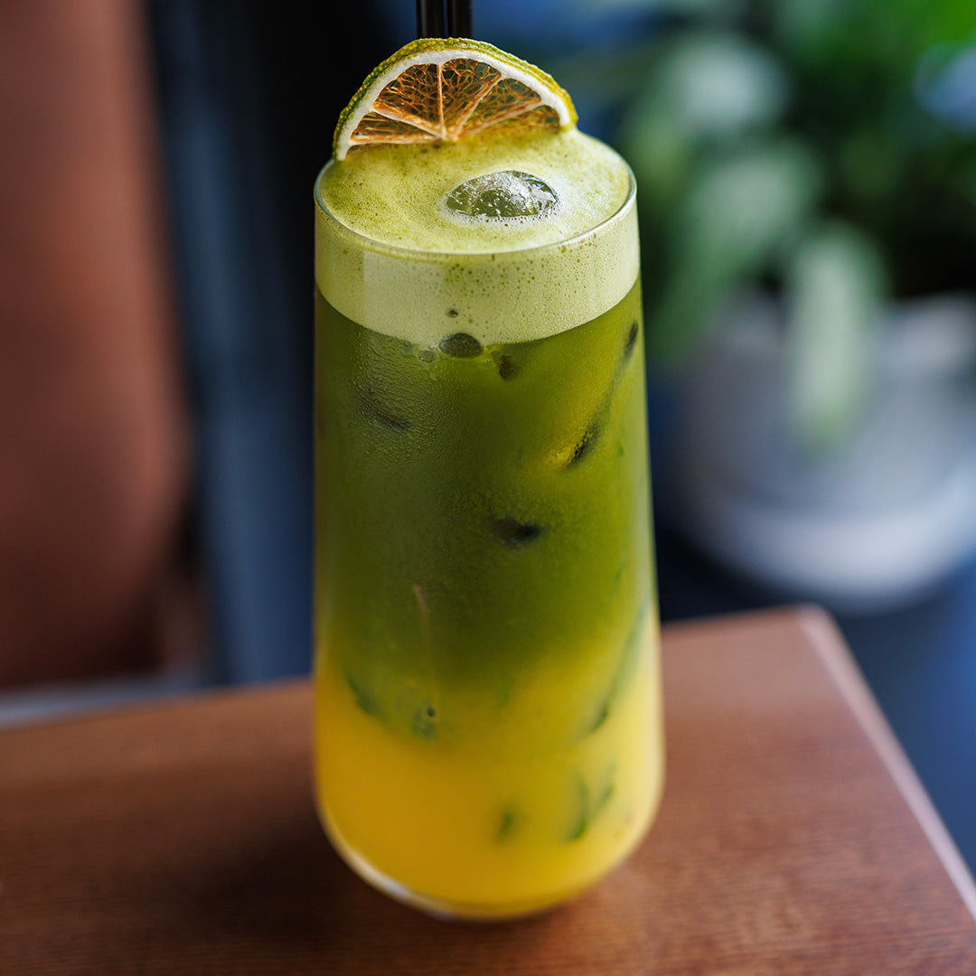 A tall clear glass filled with a layered iced drink, showing green on top and yellow on the bottom, garnished with a lemon slice on a black straw, set on a wooden table with a blurred background.