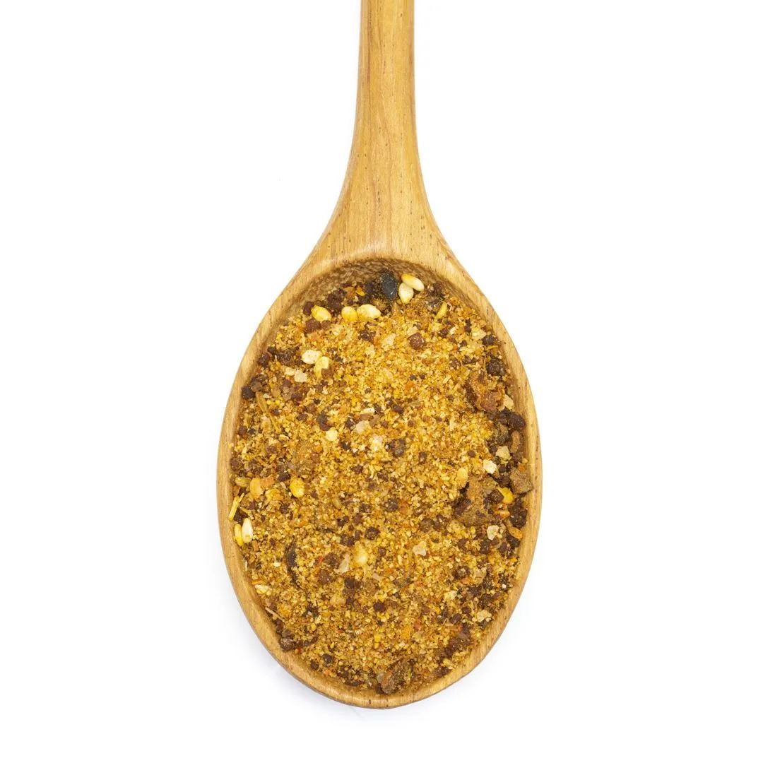 A wooden spoon filled with Mexican Mole' Spice Blend, a yellowish-brown, coarse meat seasoning with visible flakes and grains, photographed on a white background.