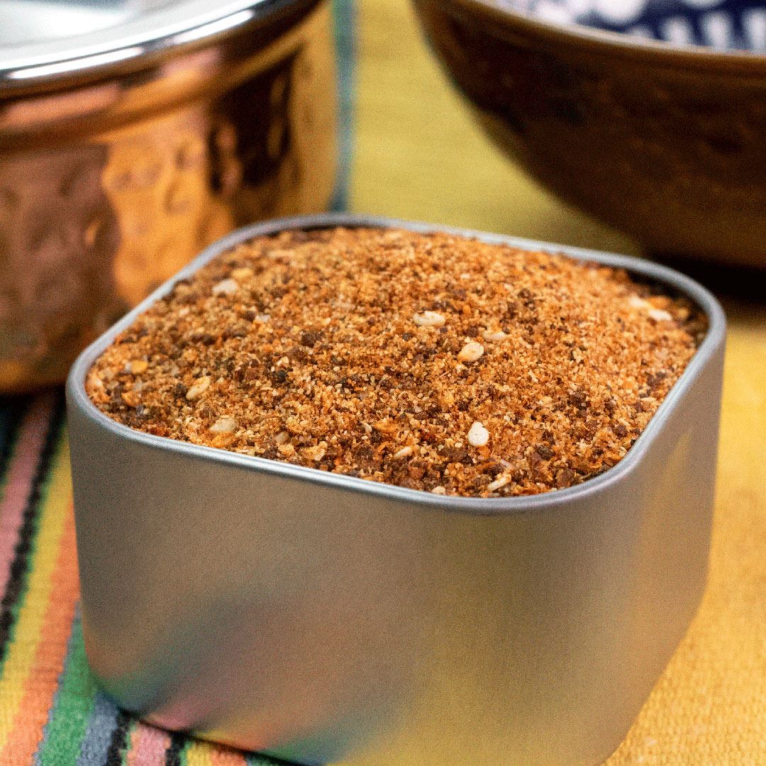 A square metal container of Mexican Mole' Spice Blend rests on a colorful striped cloth, with two bowls in the background.