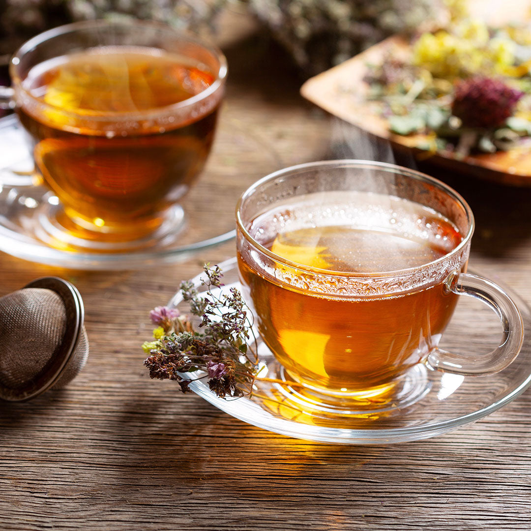 Two cups of tea on a wooden table with dried herbs and a teapot.