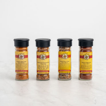 The Must-Have BBQ Rubs 4-Pack Gift Set features four glass spice jars with yellow and red labels lined up on a white surface, showcasing flavors like Carolina, Hickory, and Smokehouse Spice Blend.