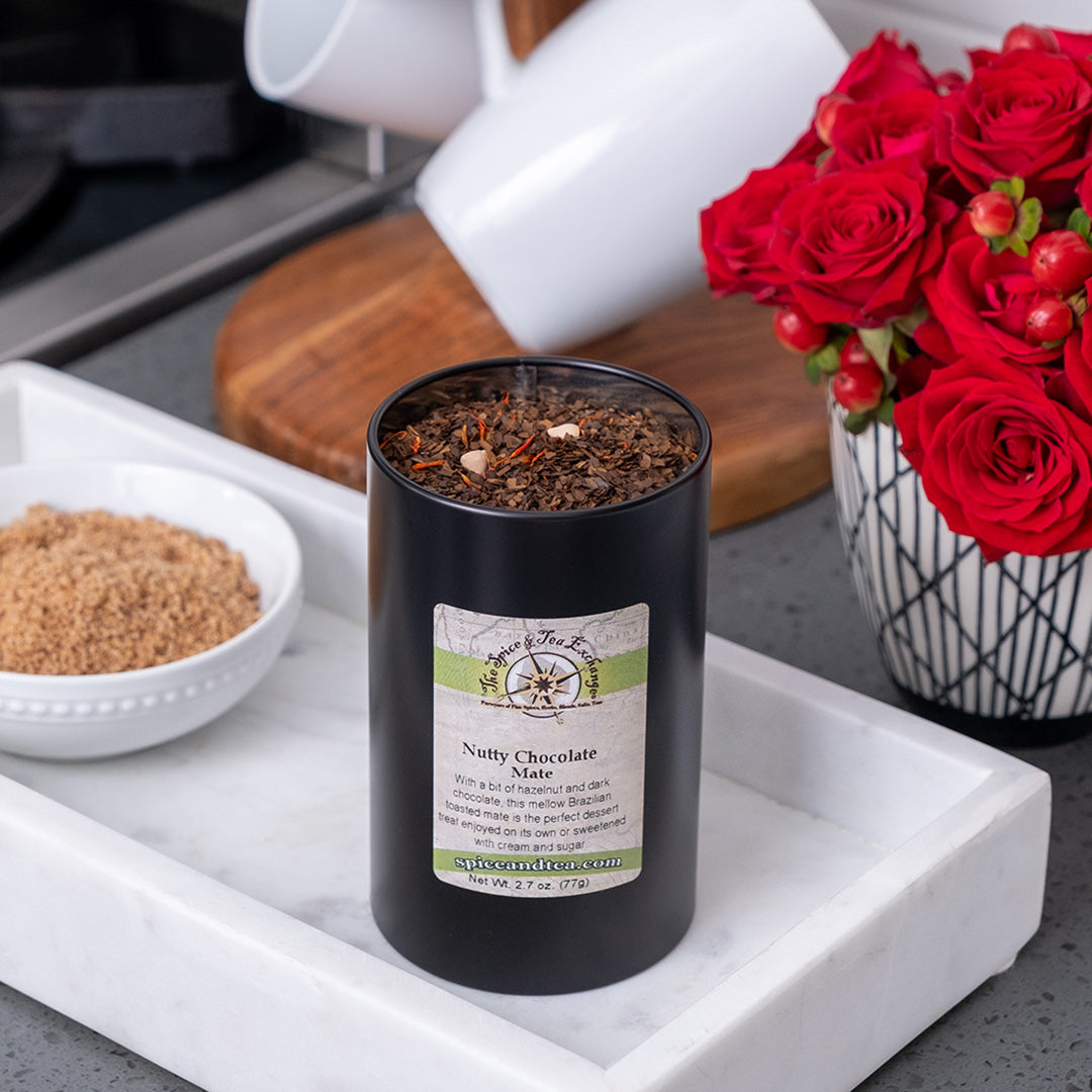 A black tin of Nutty Chocolate Mate loose leaf tea, featuring Brazilian toasted mate and hazelnut notes, rests on a white tray next to a bowl of brown sugar and red roses on a kitchen counter. White mugs are in the background.