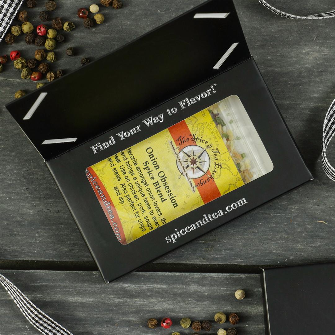 A box labeled "Find Your Way to Flavor!" holds a jar of Onion Obsession Spice Blend - Barter Box from The Spice & Tea Exchange—an ideal gift. Peppercorns and ribbon decorate the dark wooden surface around it.