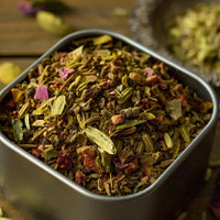 A close-up of a metal container filled with Beauty Rest Herbal Tea, a colorful blend with relaxing botanicals like fennel seeds, cardamom pods, dried rose petals, and valerian root. A small dish of extra spices appears in the background.