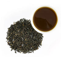 A pile of loose leaf Blueberry Black Tea sits beside a cup of dark brown brewed tea on a white background.