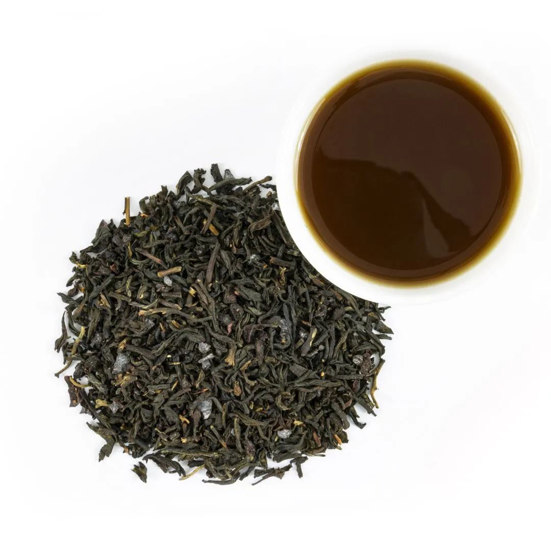 A pile of loose leaf Blueberry Black Tea sits beside a cup of dark brown brewed tea on a white background.