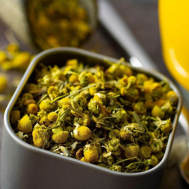 A close-up of Daily Defense Herbal Tea in a square container, filled with dried chamomile flowers and stems in yellow and green hues. The blurred background hints at more chamomile—ideal for brewing a restorative cup.