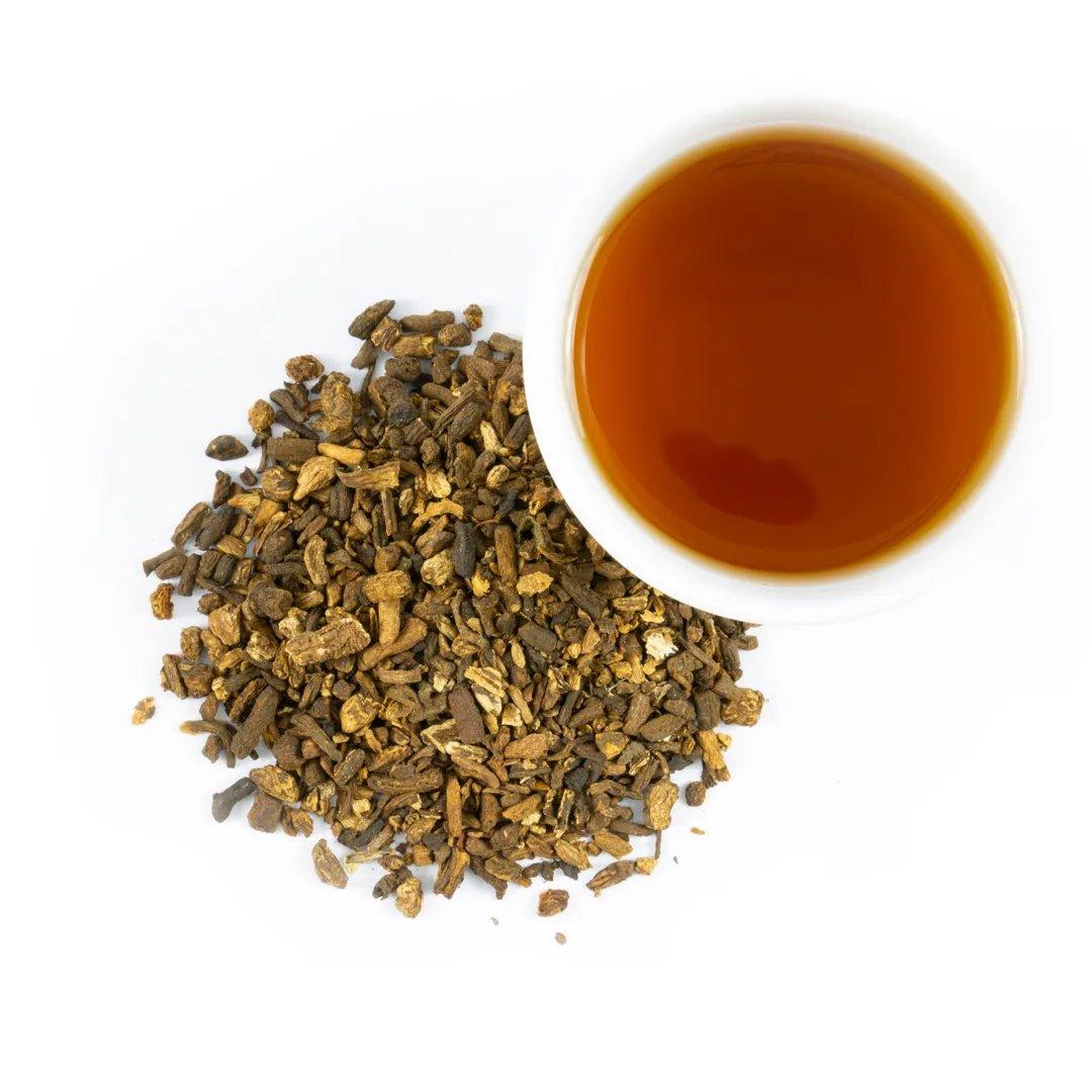 A cup of Dandelion Root - Roasted herbal tea sits beside a pile of loose, dried roots and herbs on a white background, offering a perfect coffee alternative.