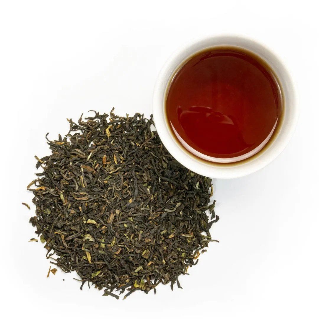 A pile of Organic Darjeeling Tea rests beside a white cup filled with dark amber tea on a white background.