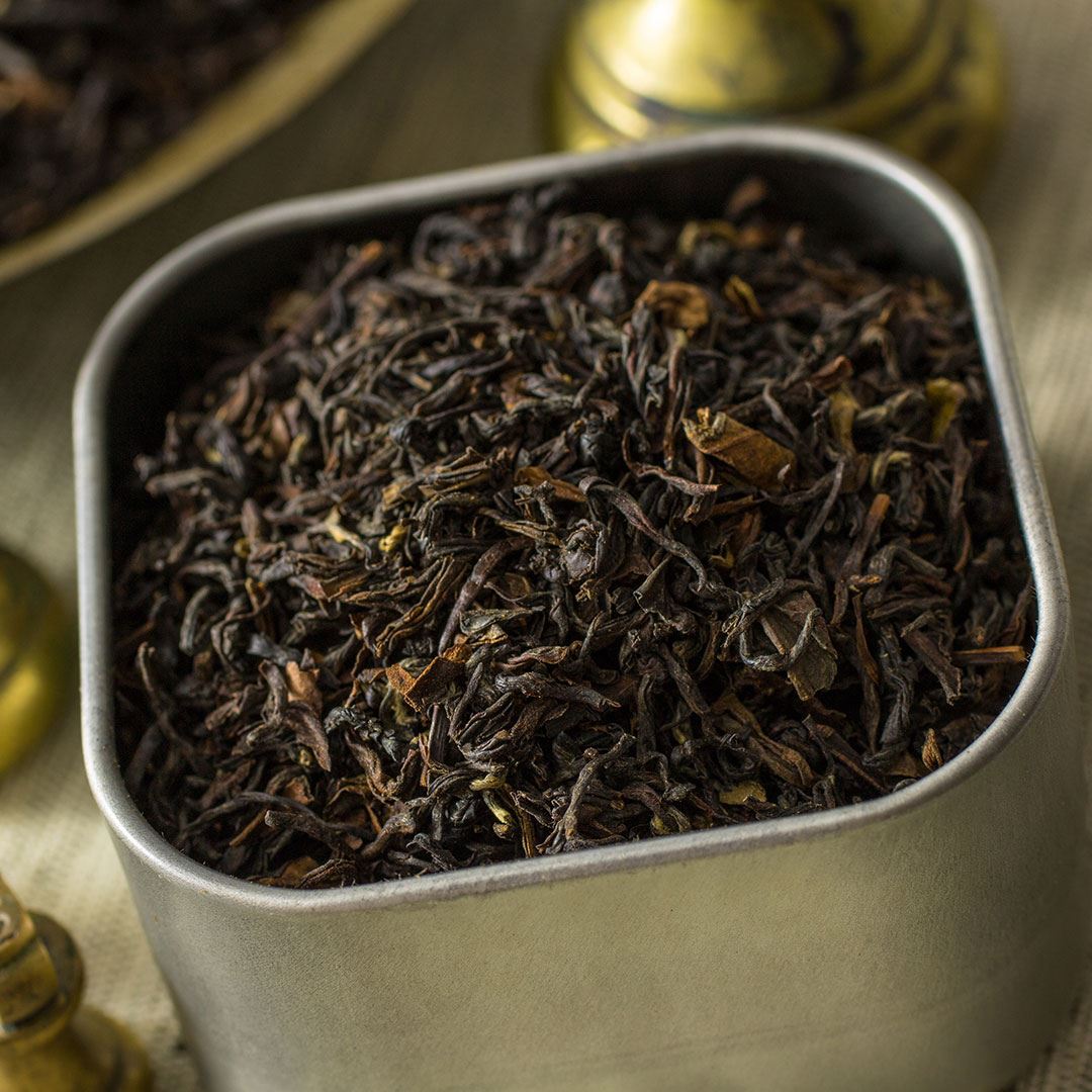 A close-up of Organic Darjeeling Tea leaves in a square metal container, with brass accents and fabric in the background.