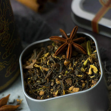 A close-up of Emperor's Chai Tea shows loose leaf black tea blended with masala spices, topped with star anise. Other visible spices and tea leaves sit in a metal tin, with part of a decorative container and tin lid in the background.