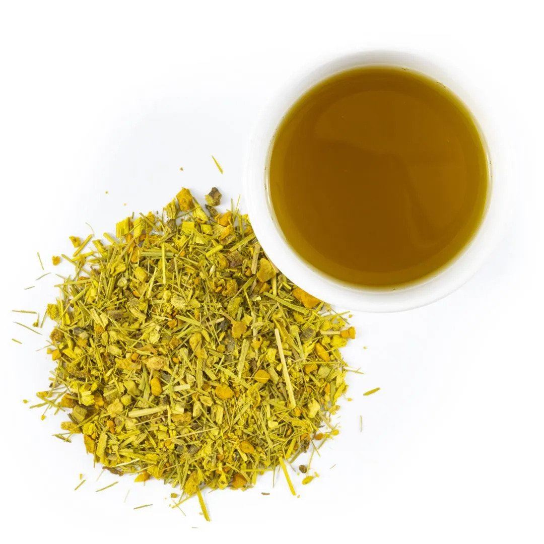 A white cup of Ginger Turmeric Herbal Tea sits beside a pile of loose, dried yellow and green herbs on a white background.