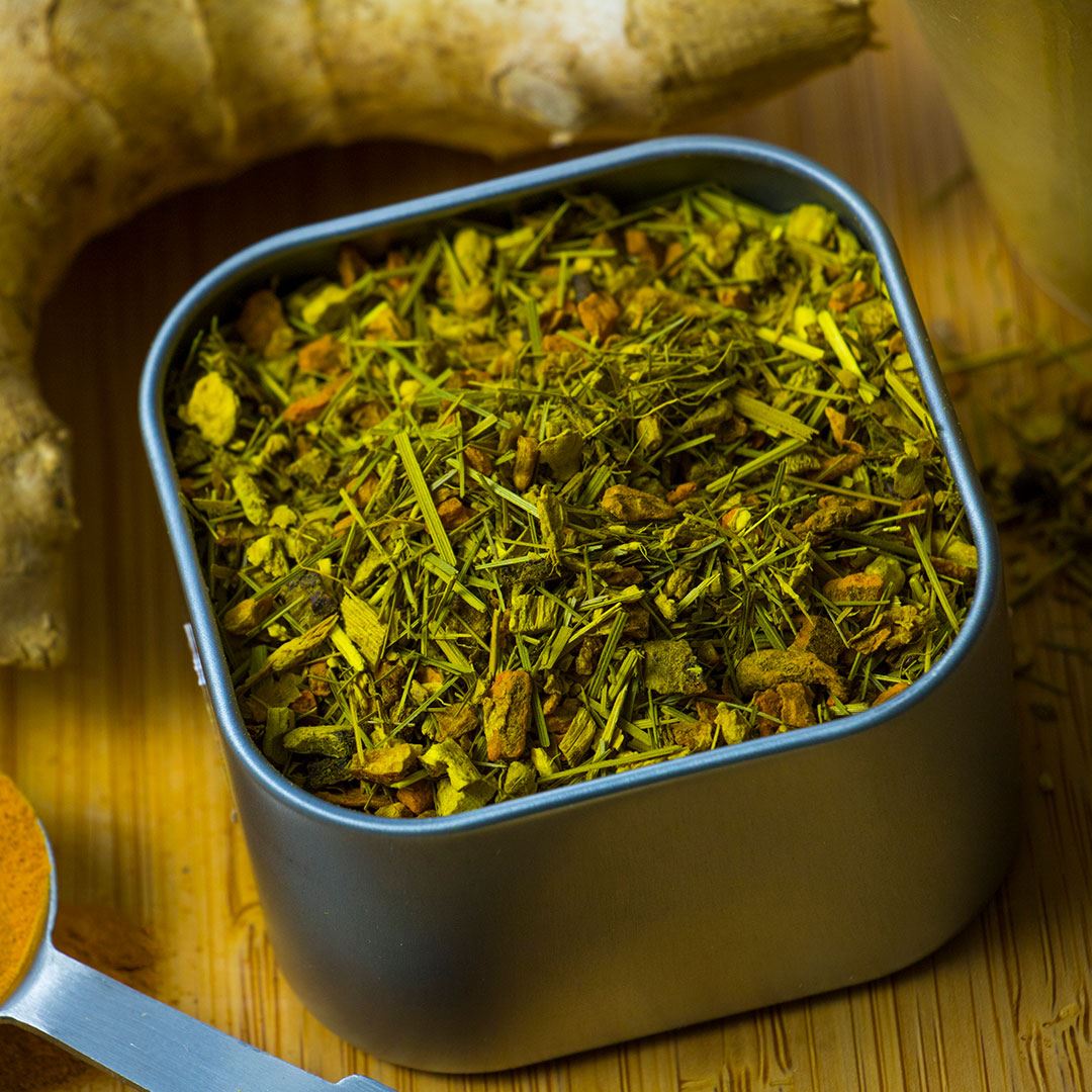 A square metal tin of Ginger Turmeric Herbal Tea with loose green and yellow leaves sits on a wooden surface, surrounded by ginger root, turmeric pieces, and a spoon.