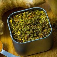 A square metal tin of Ginger Turmeric Herbal Tea with loose green and yellow leaves sits on a wooden surface, surrounded by ginger root, turmeric pieces, and a spoon.