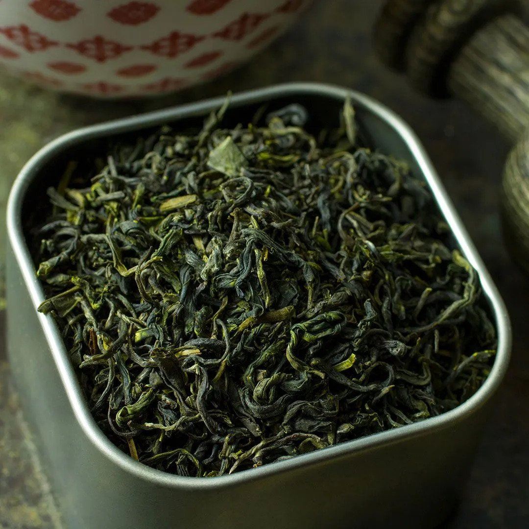 A square metal container of Jade Cloud Tea, filled with loose-leaf high-elevation green tea from Hubei, China. A patterned bowl and other tea accessories are softly blurred in the background.