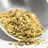 Close-up of a metal spoon holding Licorice Root - Cut & Sifted, with dried yellowish-brown pieces scattered on a white surface—commonly used to soothe sore throats and aid digestion.