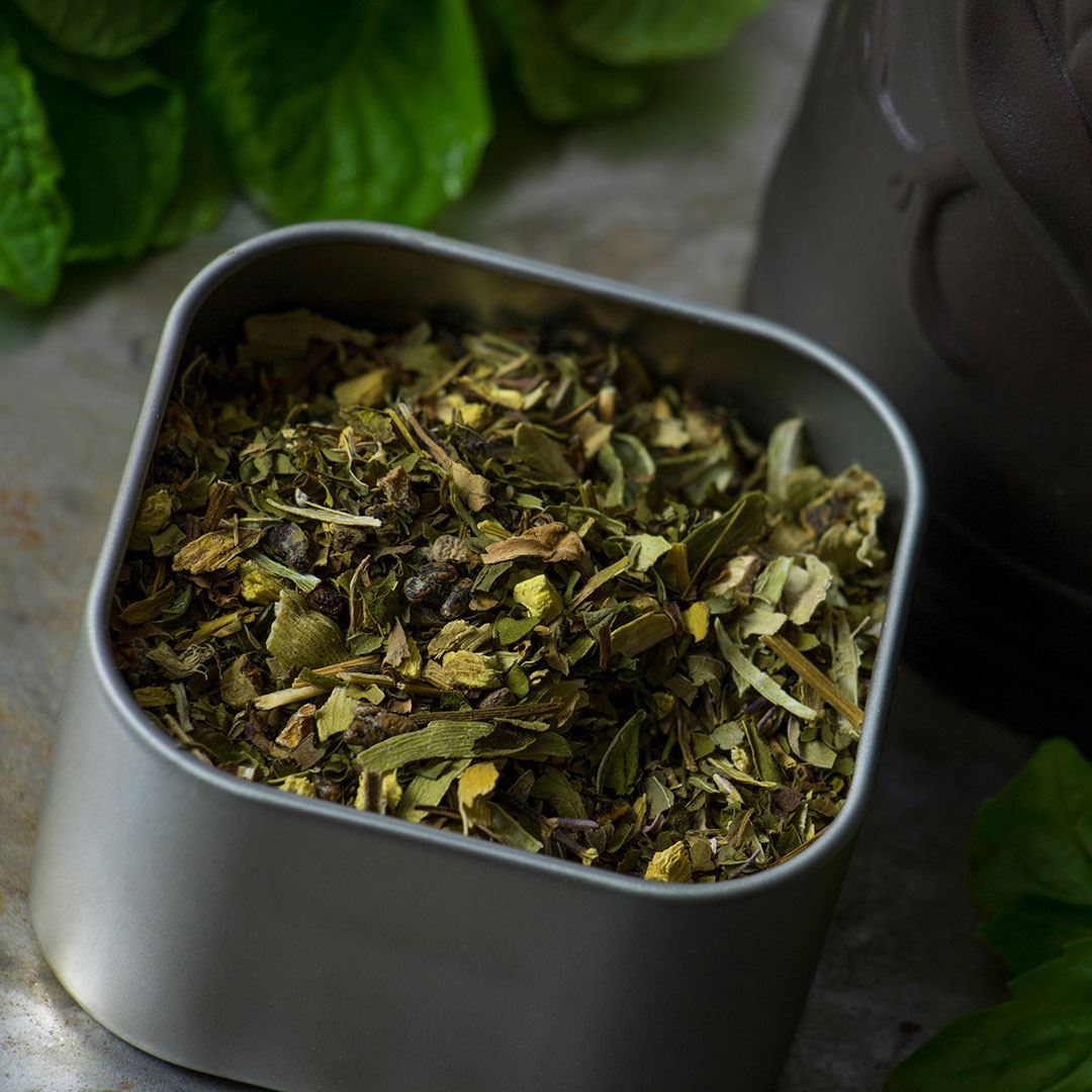 A square metal tin of Mint Basil Herbal Tea, filled with a blend of dried green herbs, rests on a stone surface, accompanied by fresh green leaves and part of a dark container in the background.
