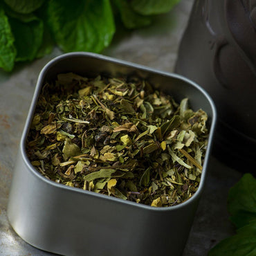 A square metal tin of Mint Basil Herbal Tea, filled with a blend of dried green herbs, rests on a stone surface, accompanied by fresh green leaves and part of a dark container in the background.