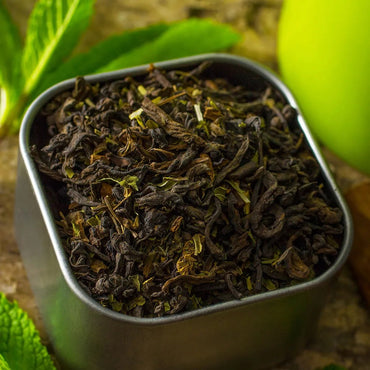 A close-up of Mint-Chilla Chai-Nilla Tea loose leaves in a square metal container, with fresh mint leaves and a green object softly blurred in the background.