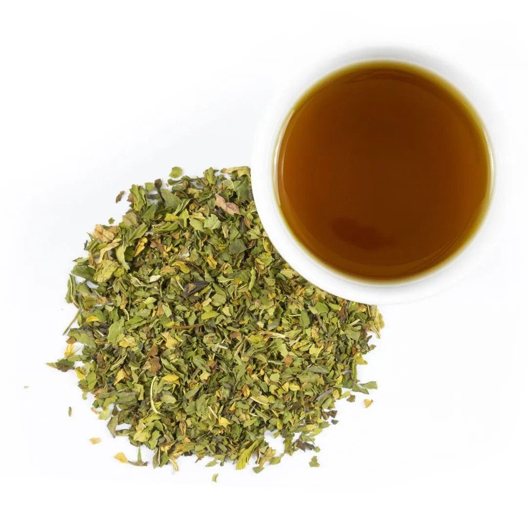 A cup of brown tea, highlighted by Organic Peppermint, sits on a white background beside a loose pile of dried green tea leaves for a refreshing touch.