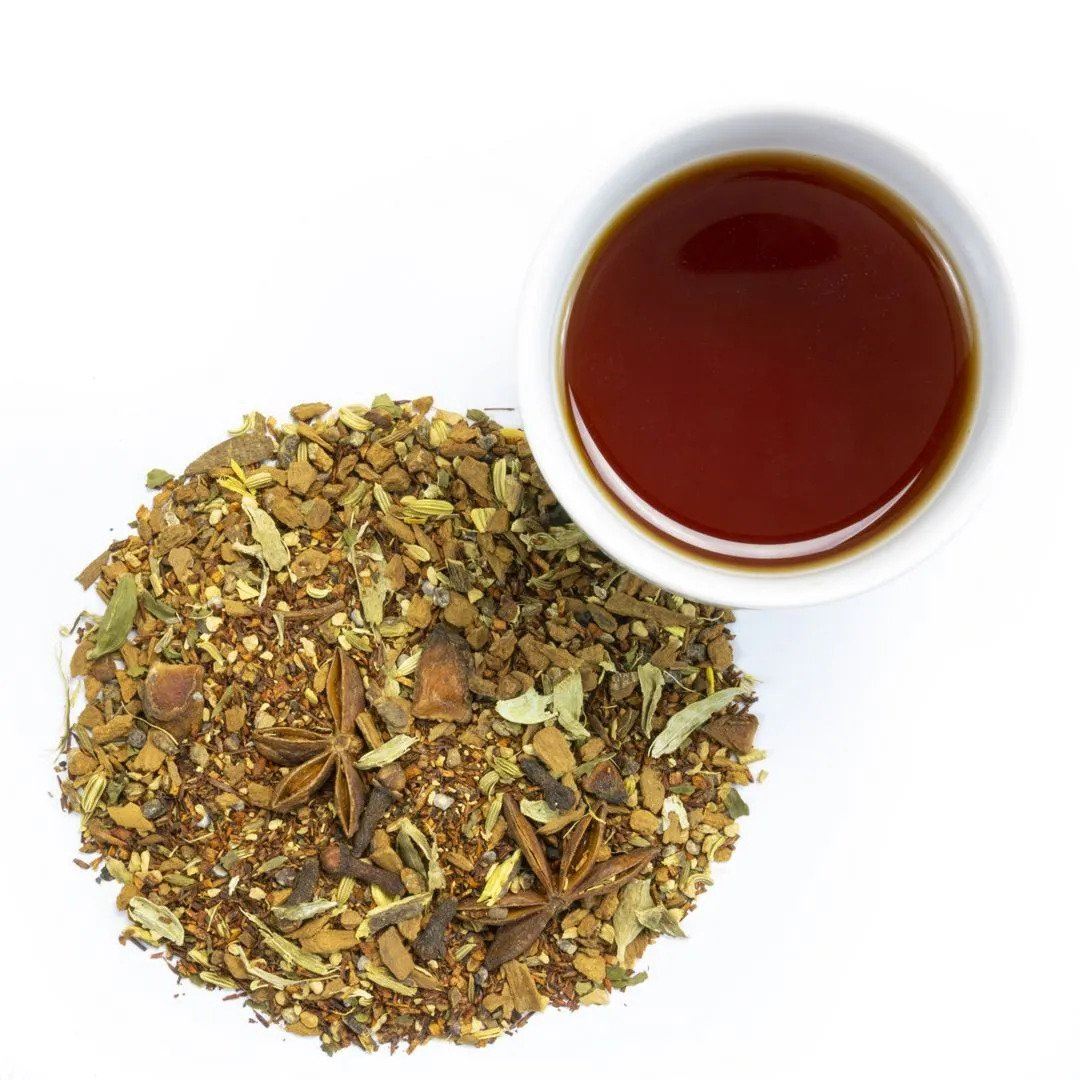 A cup of Rooibos Chai Herbal Tea, surrounded by herbs, suggests its sweet, spiced flavor.