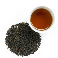 Loose, dark Ruby Oolong Tea leaves rest beside a white cup of amber-hued brewed tea on a pristine white background, suggesting the rich, complex flavor within.