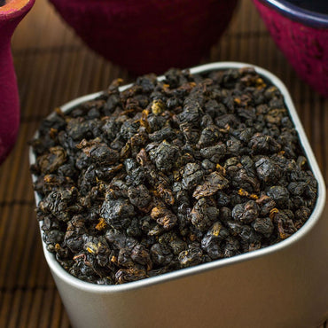 A square metal container of Ruby Oolong Tea sits on a bamboo mat, with glimpses of colorful cups in the background, suggesting the complex flavors of this distinctive oolong.