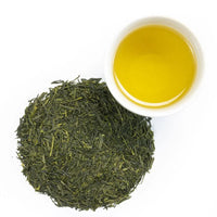 A pile of Sencha Superior Tea leaves sits beside a white cup filled with vibrant yellow-green tea, highlighting fresh grassy notes and a smooth body, all set against a crisp white background.