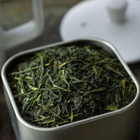 A close-up of loose Sencha Superior Tea leaves in a square metal container, known for their fresh grassy notes, with a white teapot lid partially visible in the background.