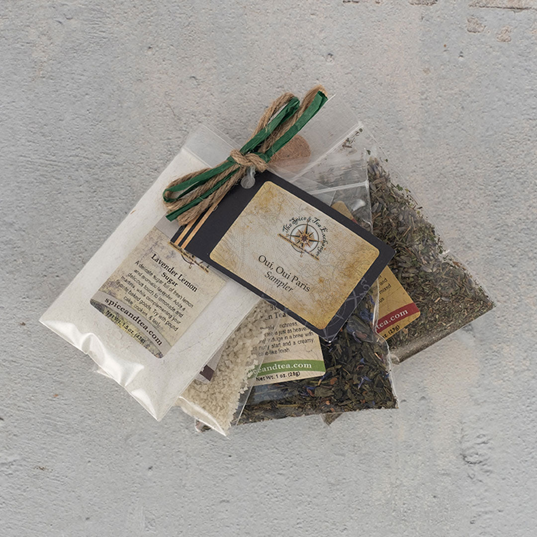 Three small clear bags filled with French seasonings and grains, tied with twine and a green ribbon, rest on a light gray surface. A rustic tag labels them as the "Oui, Oui Paris Sampler.