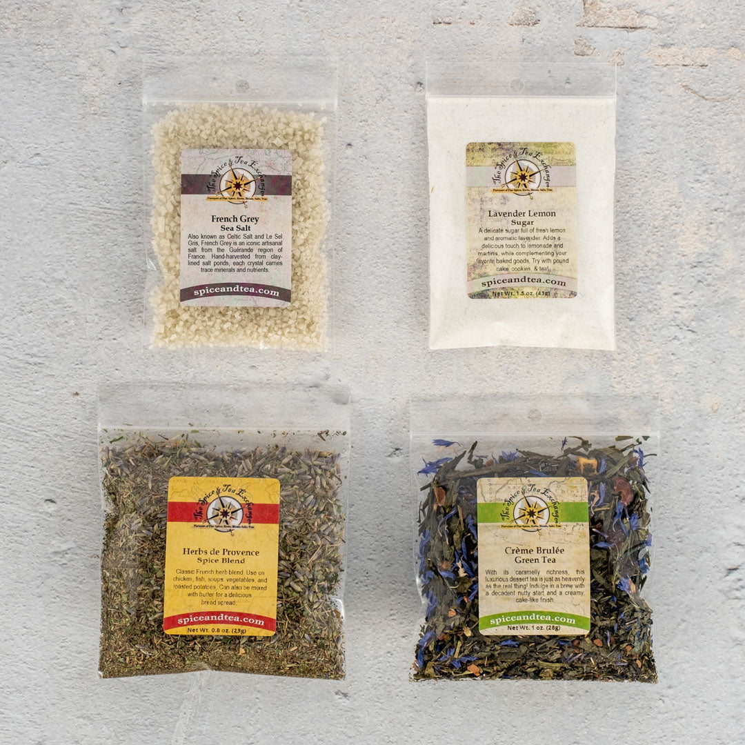 Four clear packets rest on a light surface, each labeled with colorful tags. The Oui, Oui Paris Sampler includes French Grey Sea Salt, Lavender Lemon sugar, Herbs de Provence spice blend, and Crème Brûlée green tea.