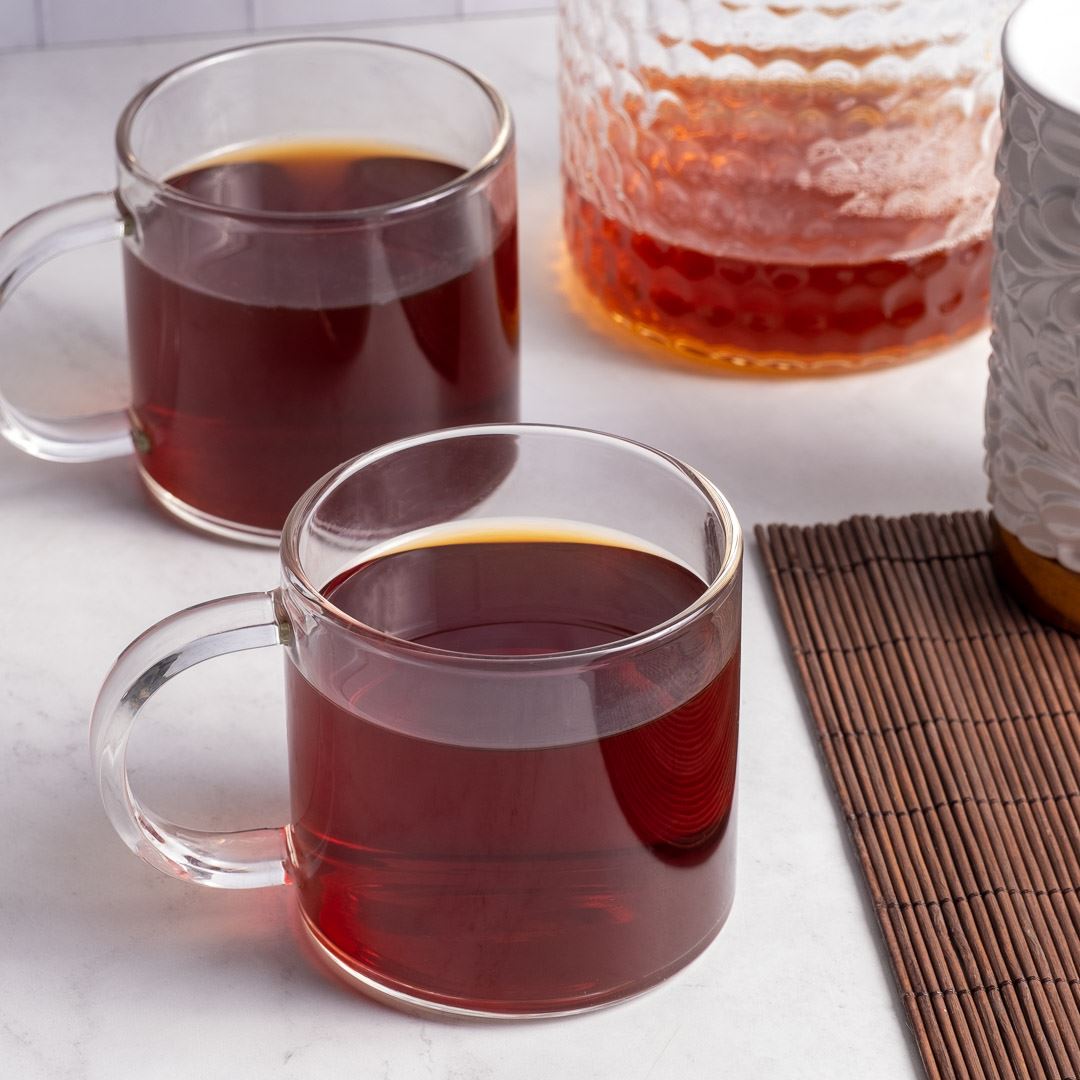 Two clear glass mugs filled with Peach On The Beach Tea sit on a white surface. A textured glass jug holds extra tea, and part of a white decorative container rests nearby next to a brown bamboo mat.