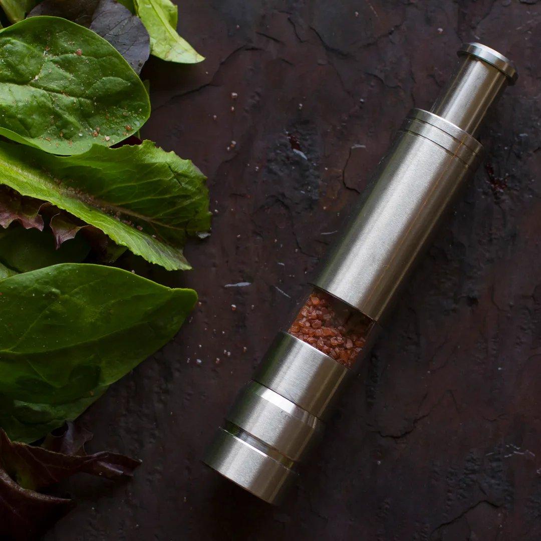 A Pump & Grind Salt & Pepper Mill filled with pink salt rests on a dark stone surface beside fresh green and purple salad leaves.