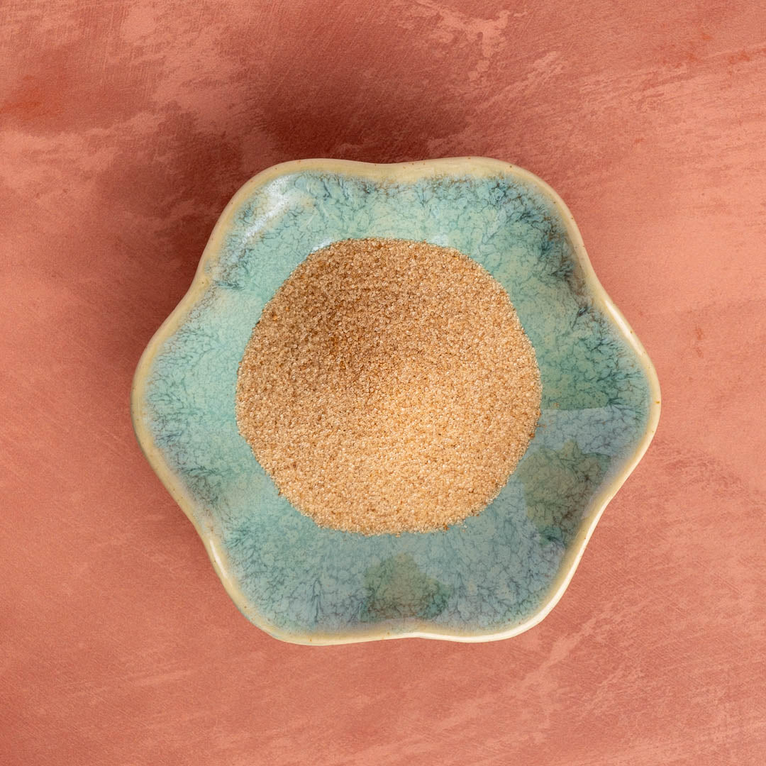 A small, round, blue-green ceramic dish holds a mound of light brown Pumpkin Spice Monk Fruit Sweetener, a natural sweetener, set atop a textured reddish-brown surface.