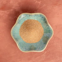 A small, round, blue-green ceramic dish holds a mound of light brown Pumpkin Spice Monk Fruit Sweetener, a natural sweetener, set atop a textured reddish-brown surface.