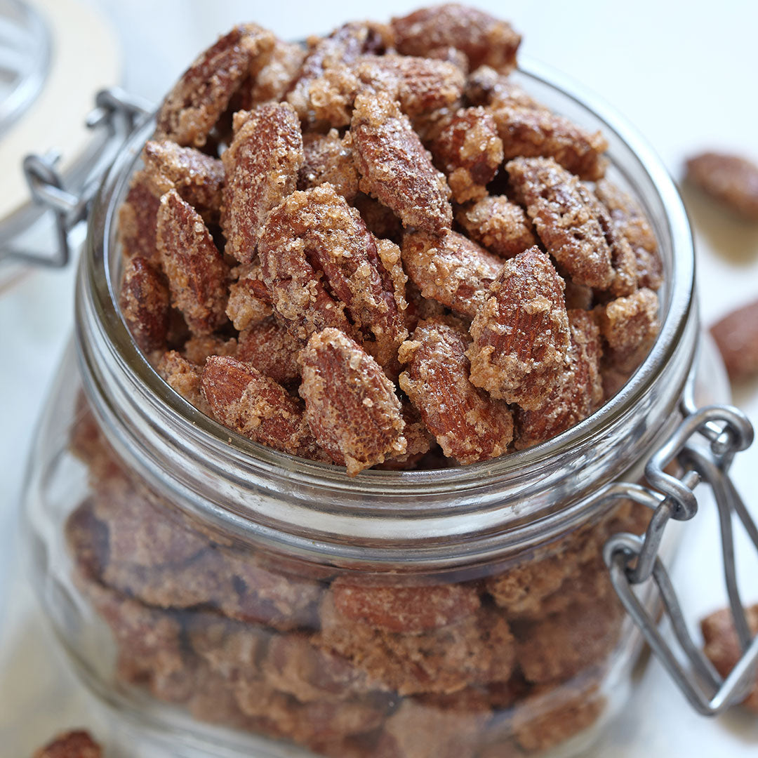 A glass jar brimming with candied pecans coated in Pumpkin Spice Monk Fruit Sweetener, with a few spilling out onto the white surface, showcases their irresistible appeal.