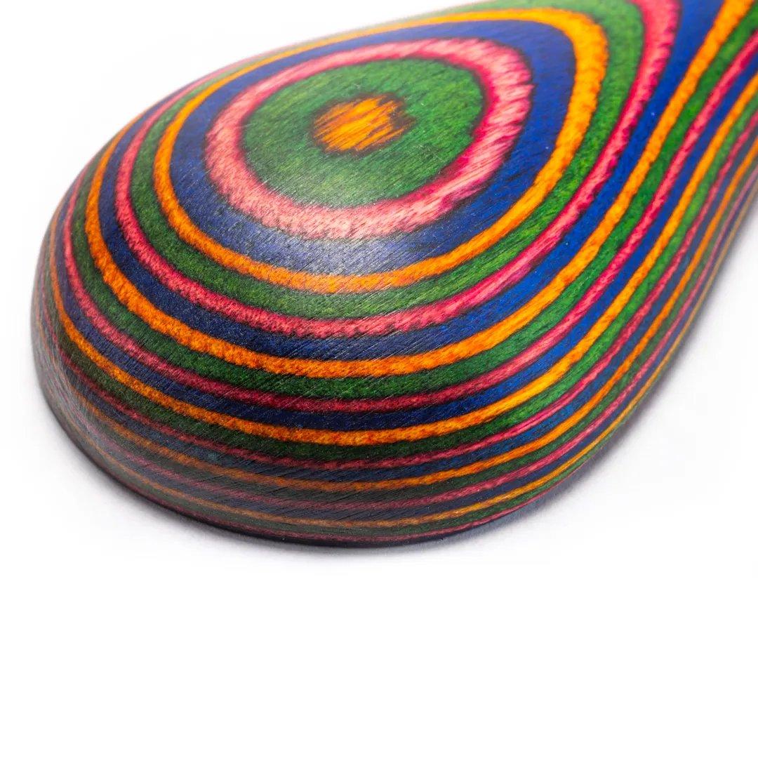 A close-up of the Rainbow Double Ended Spoon, crafted from smooth laminated Pakkawood with vibrant concentric stripes in green, pink, blue, orange, and yellow on a white background.